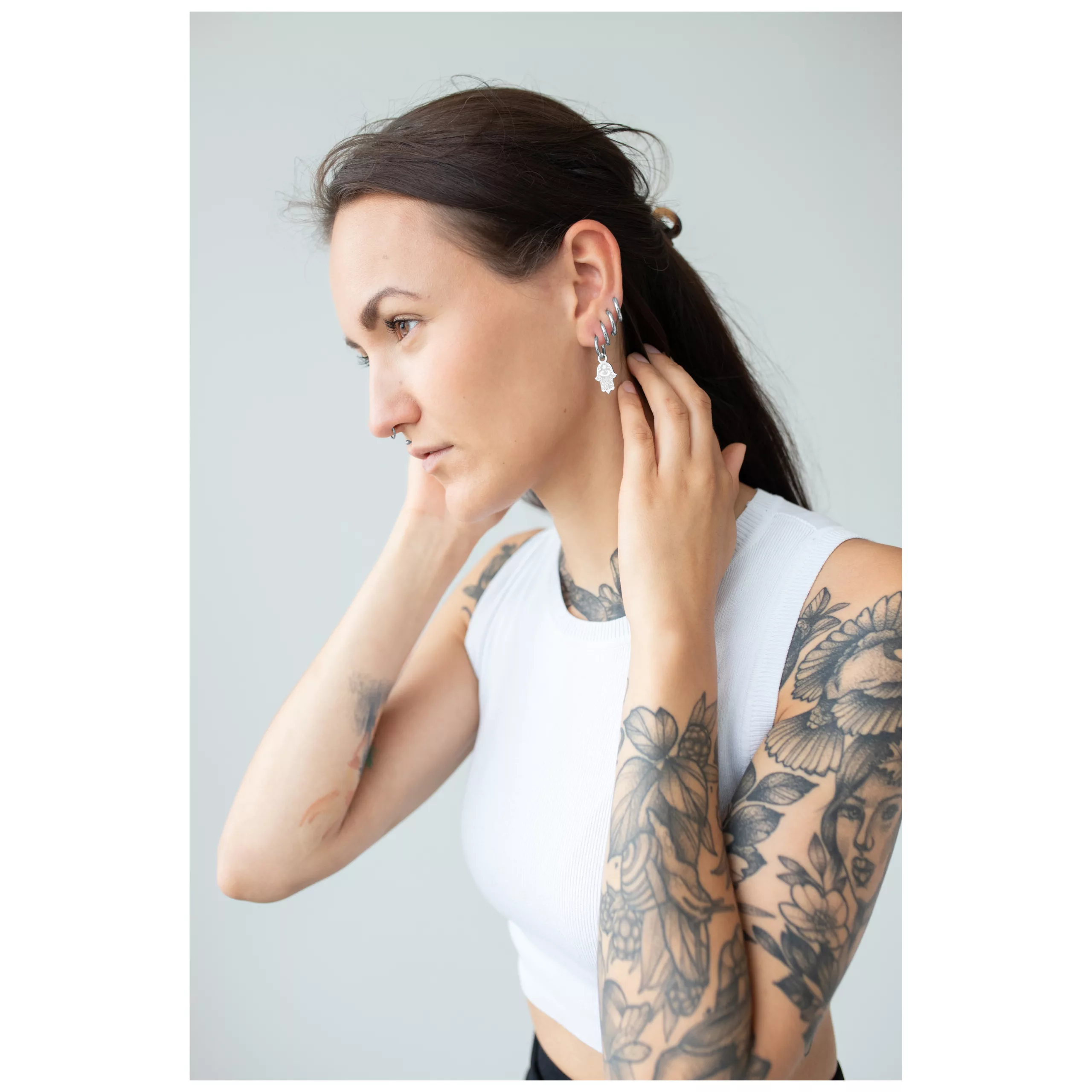 Little Hamsa Hand Hoops am Ohr getragen Frau mit Tätowierungen trägt Hamsa Hand Ohrringe, weißes Top, grauer Hintergrund