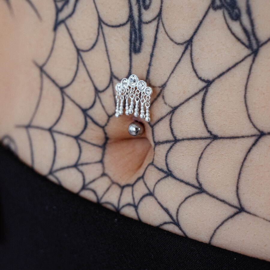 Silbernes Bananabell-Piercing mit fünf hängenden Kristallen in einem Bauchnabel, umgeben von einem Spinnennetz-Tattoo